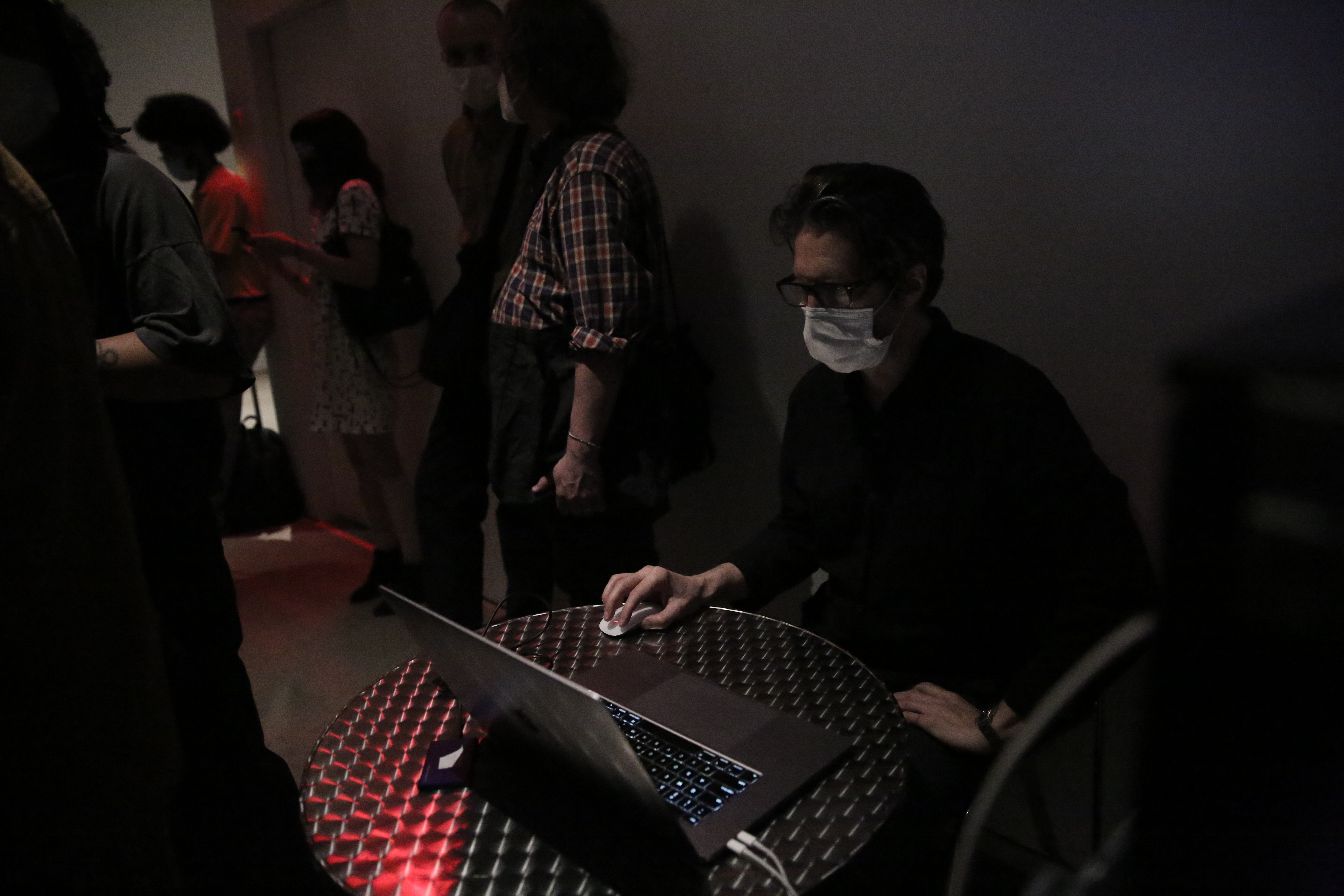 Several figures stand along a wall. One figure, seated, uses a computer at a small round table.