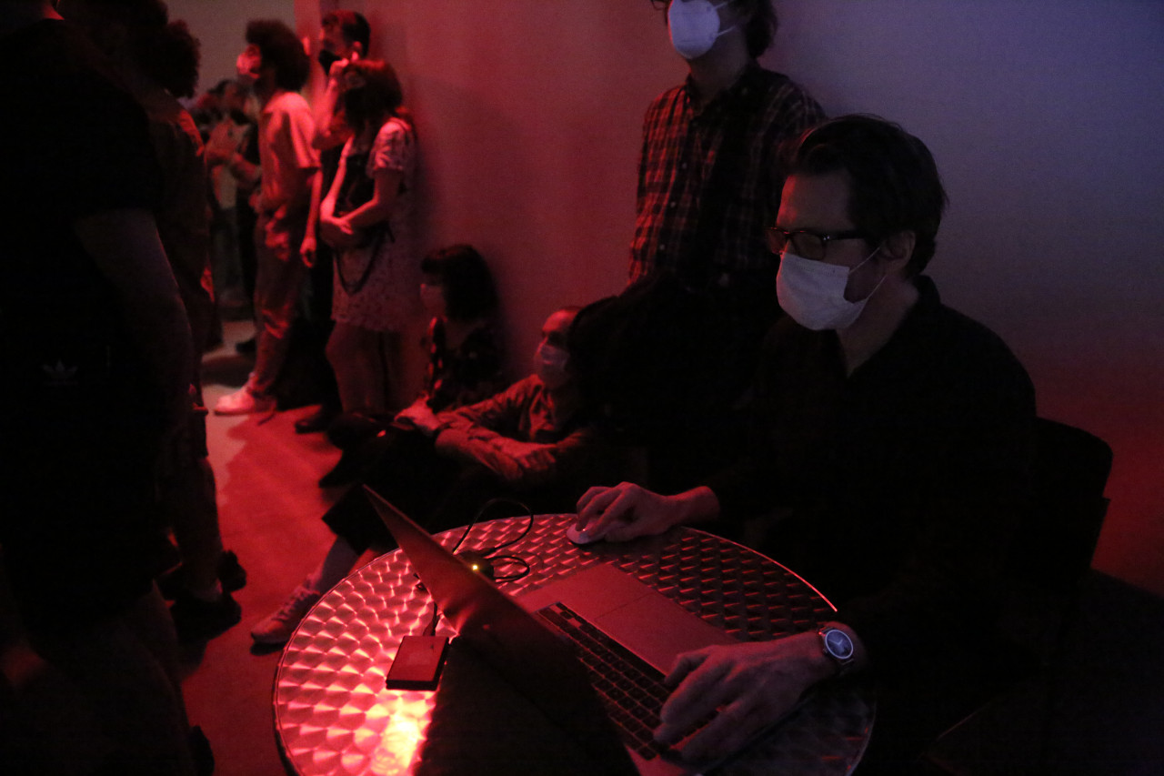 Several figures stand along a wall illuminated by red light. One figure, seated, uses a computer at a small round table.