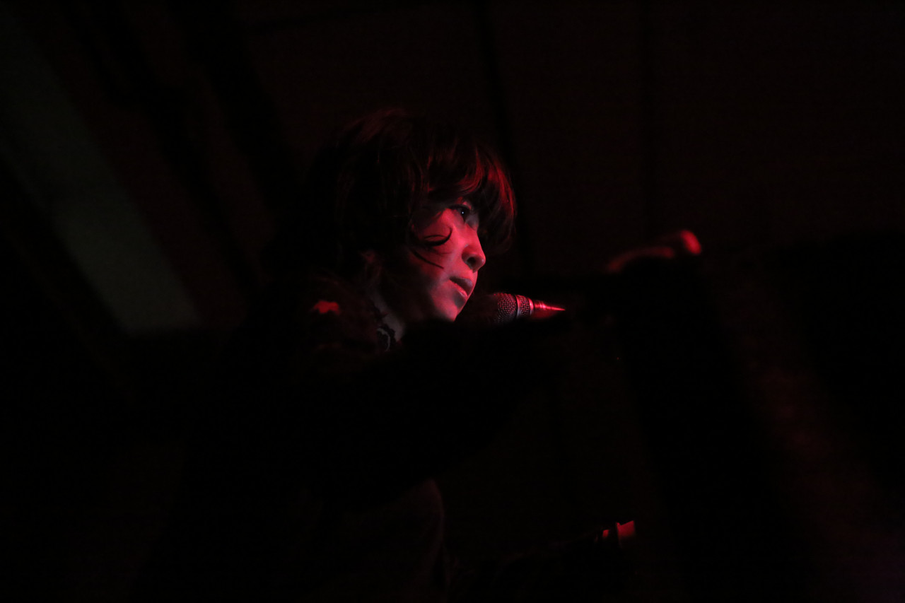 A woman with dim red lights on her face in a dark environment stands and talks behind a microphone.