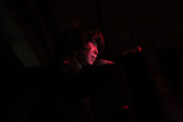 A woman with dim red lights on her face in a dark environment stands and talks behind a microphone.