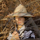 A color photograph of a figure in a pointed hat with a wide brim, wearing a black and white robe. The figure is pictured amid tree branches and vines that block their face. They hold a twisted vine in their left hand.