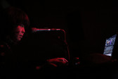 A woman stands in front of a micrphone and computer. Her face and hands are illuminated by red light.