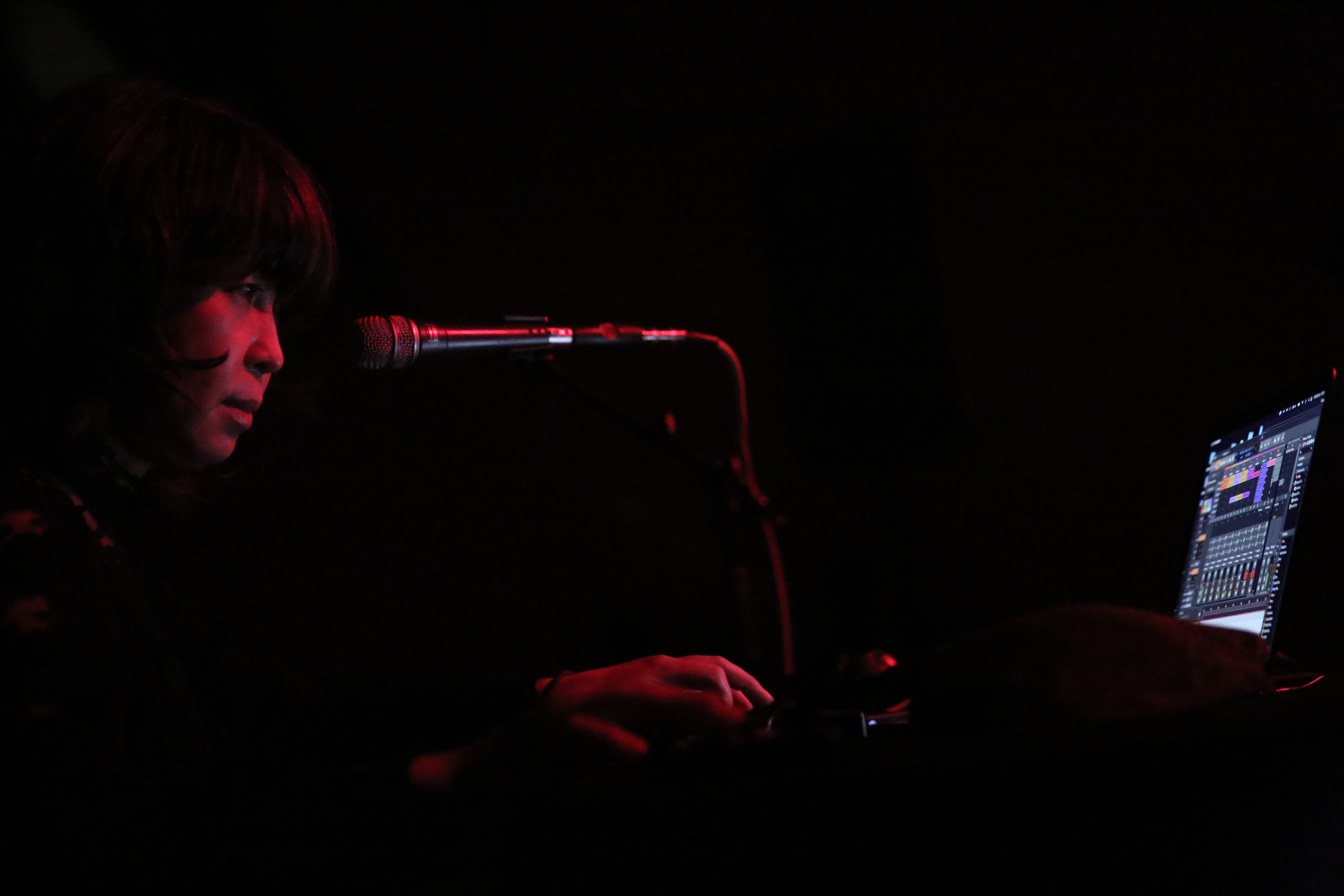 A woman stands in front of a micrphone and computer. Her face and hands are illuminated by red light.