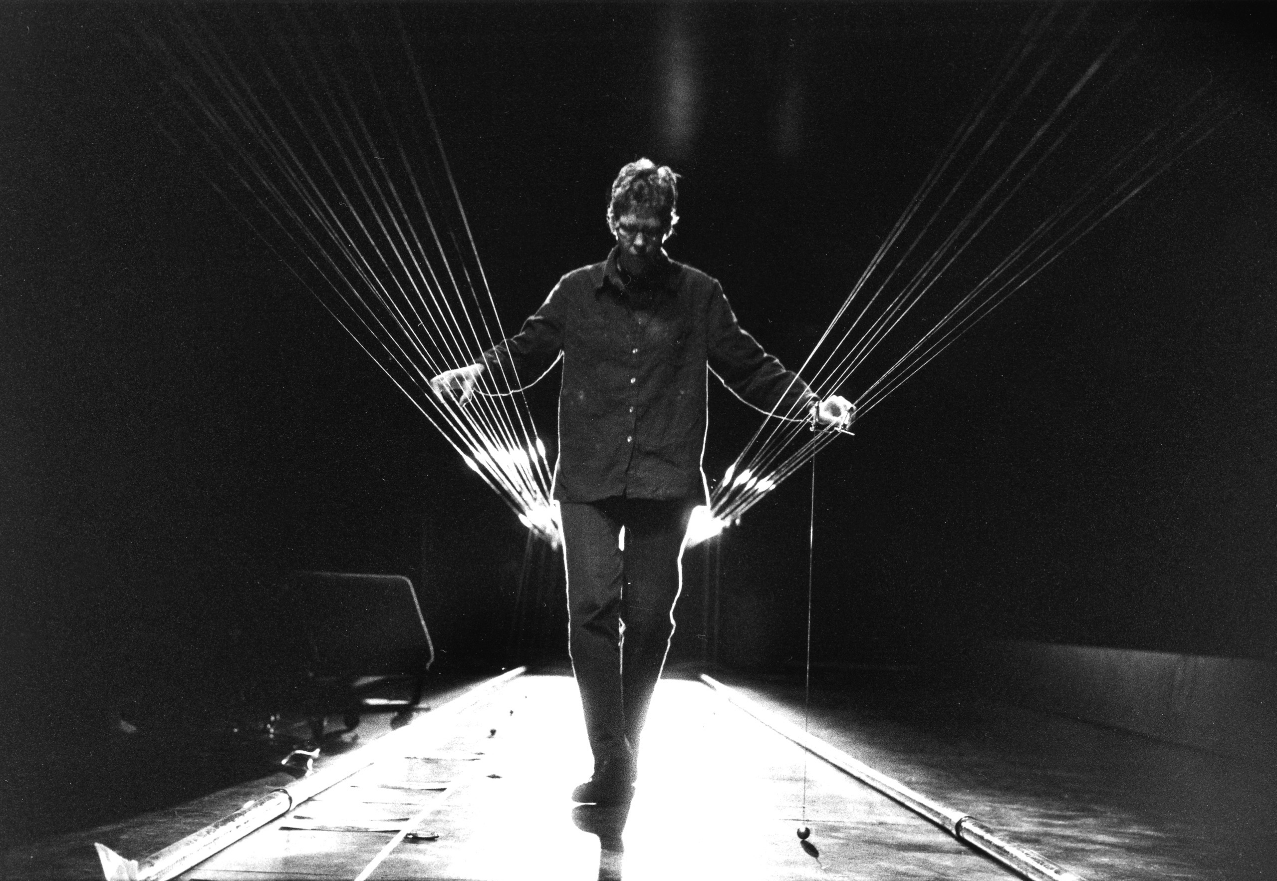 Black-and-white photograph of a figure, walking in the middle of the frame between two sets of suspended wires that run from across the room. The figures hands manipulate the wires on each side of them.