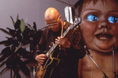 A man with a bald head wearing wire frame glasses strums an electric guitar by a set of microphones. A potted plant and a large sculpture of a baby with glowing, blue eyes are situated behind him.