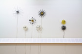 A series of vintage, abstract clocks hanging on a white wall above a long didactic text displayed on a horizontal plinth.