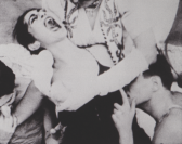 A woman screaming while two men hold her down and touch her breasts.