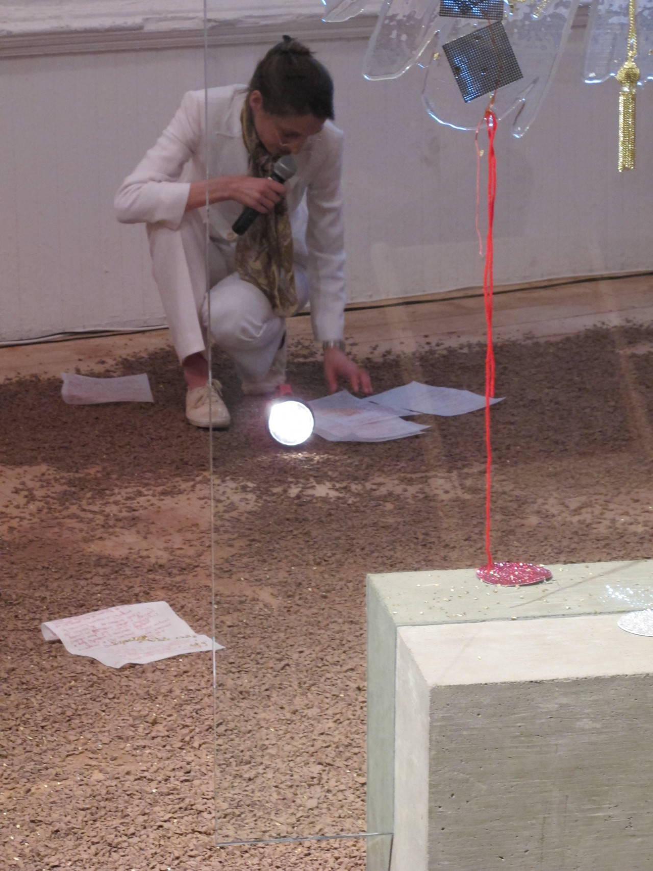 A figure in a white suit crouches on the floor of a gallery space, speaking into a microphone held in one hand while reading from a series of papers stewn about her on the ground. The ground is unevenly covered in a grainy, brown dirt-like material. A flashlight rests on the ground at her feet, pointing towards and illuminating a sculpture composed of a large glass pane wedged into the center of a square concrete block. String, golden tassles, and other sparkling ornaments hang on the glass pane.