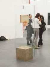 A sculpture consisting of a large rectangular pane of glass, its bottom edge wedged into the center of a square block, is installed in a white-walled gallery space. A small canvas is attached to the center of the glass pane with its wooden backing facing the viewer. String and an orange, mesh-like object hang and drip from the canvas's lower edge. Three figures are visible behind the sculpture, talking in a small cluster.