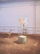 A sculpture composed of a large glass pane wedged into the center of a square concrete block sits in a gallery space. A painted canvas is installed in the pane, displaying a large number 
