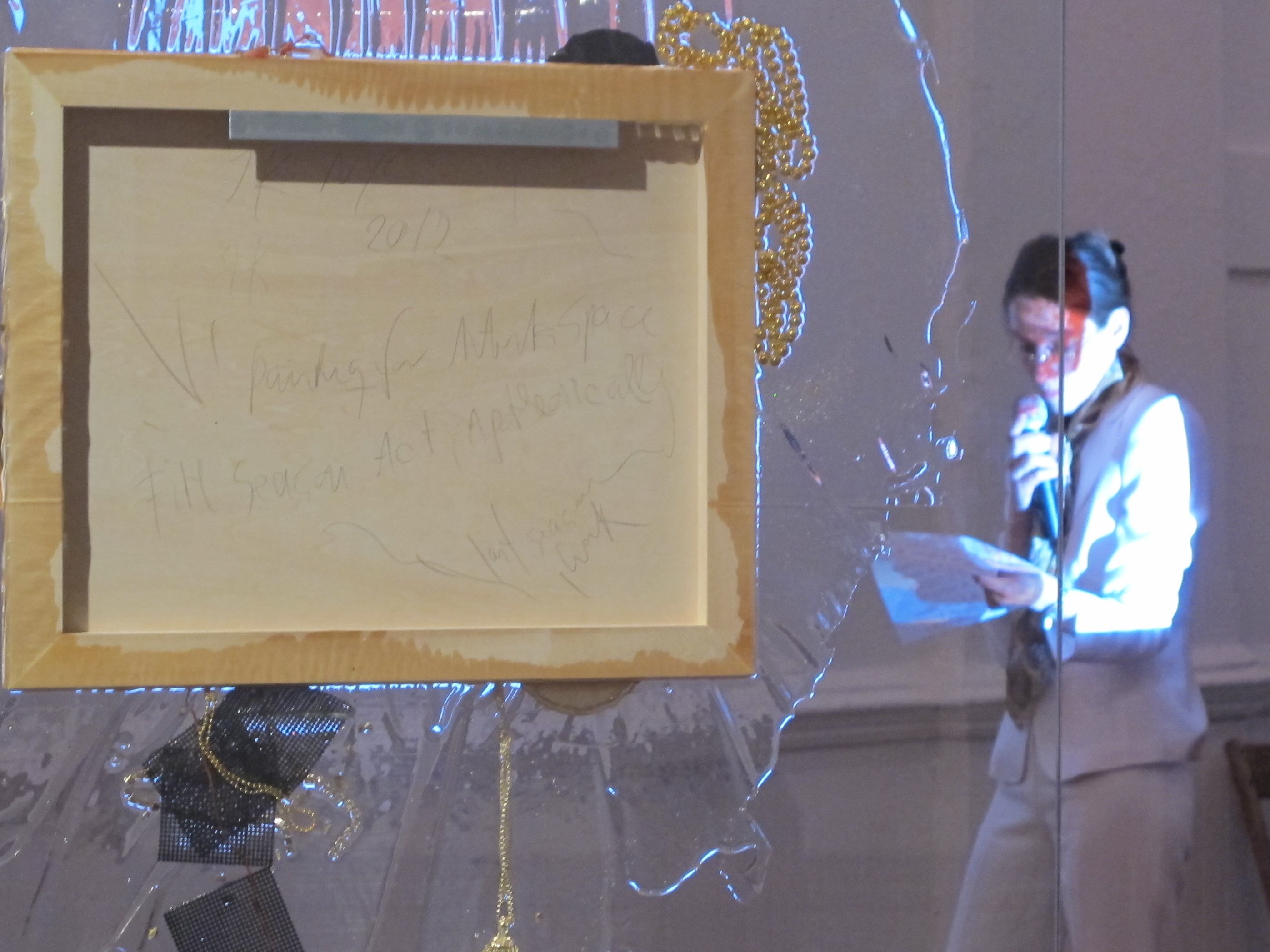 Close up detail of a sculpture composed of a large glass pane with a painted canvas installed in the pane's center. The back of the canvas faces the viewer, and faint words are penciled onto its surface. A golden tassle and necklce cluster and hang around the canvas' edge.