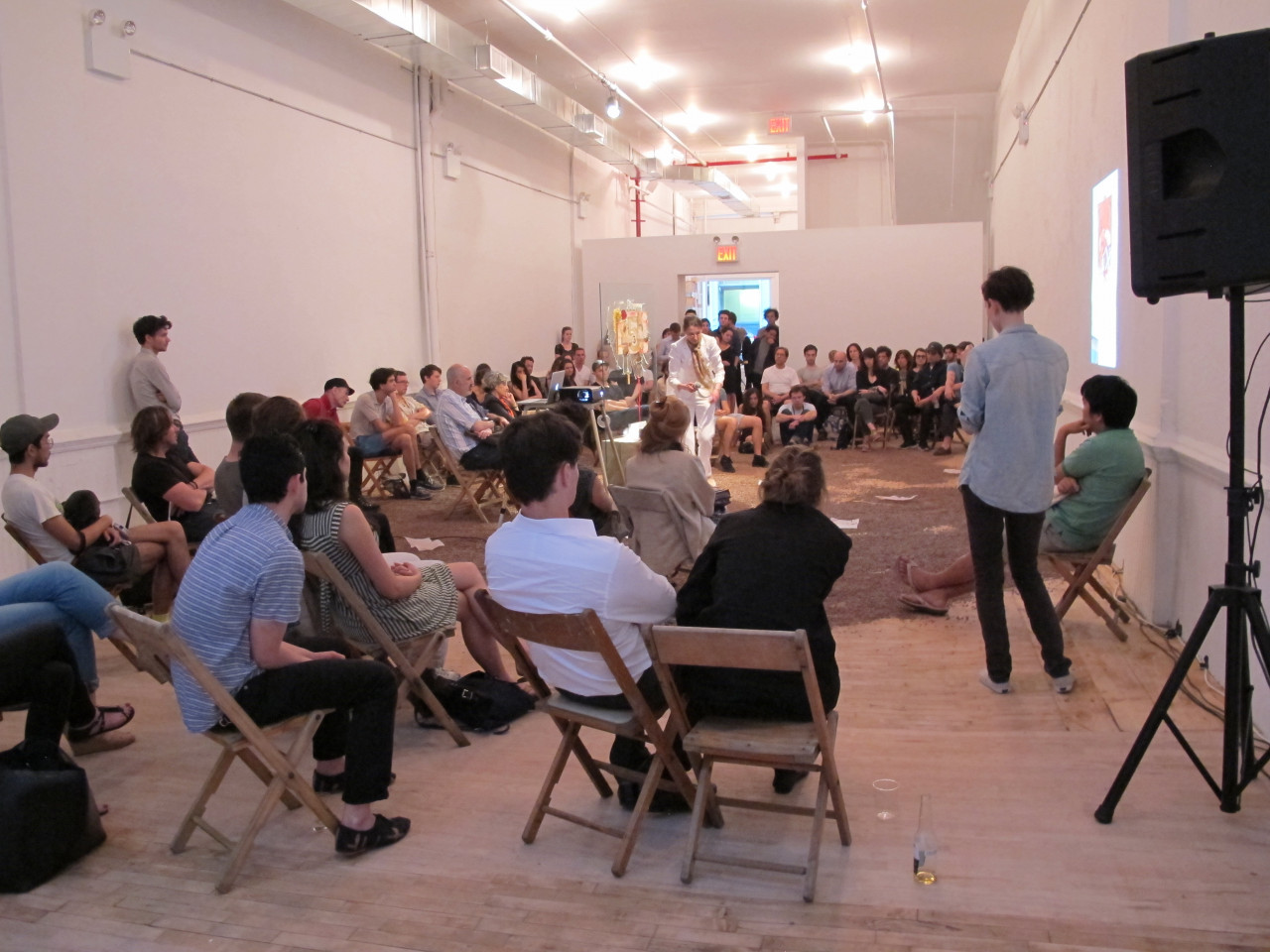 A crowd if people sit on wooden chairs in a large gallery space. The crowd forms a semi-circle around a portion of the gallery floor covered in a brown, dirt-like material. A woman in a white suit stands amonst the debris and speaks into a microphone in front of a sculpture composed of a large pane of glass.