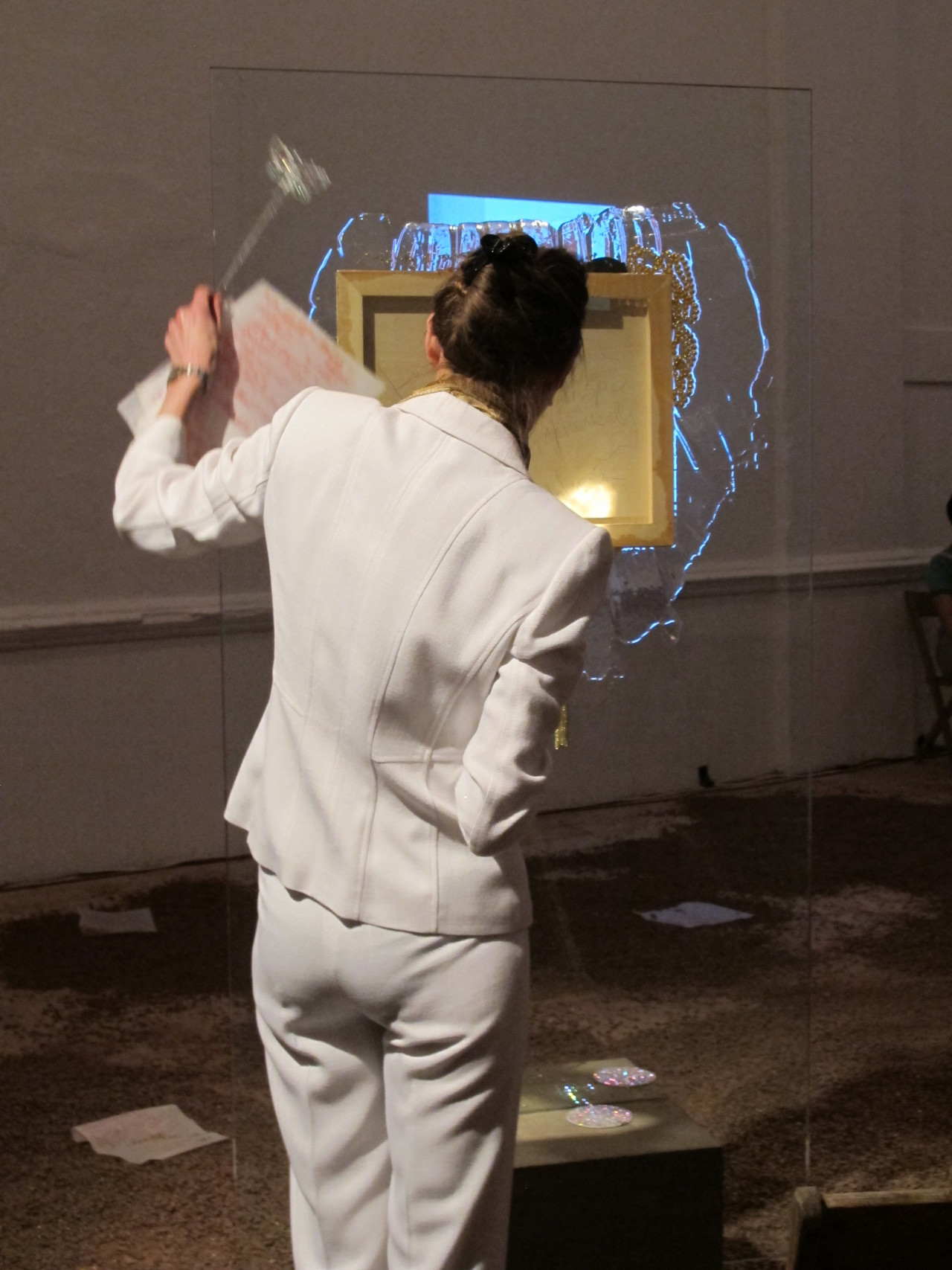A figure in a white suit—the artist Jutta Koether—stands with her back to the viewer in front of a sculpture composed of a large glass pane with a painted canvas installed in the panel