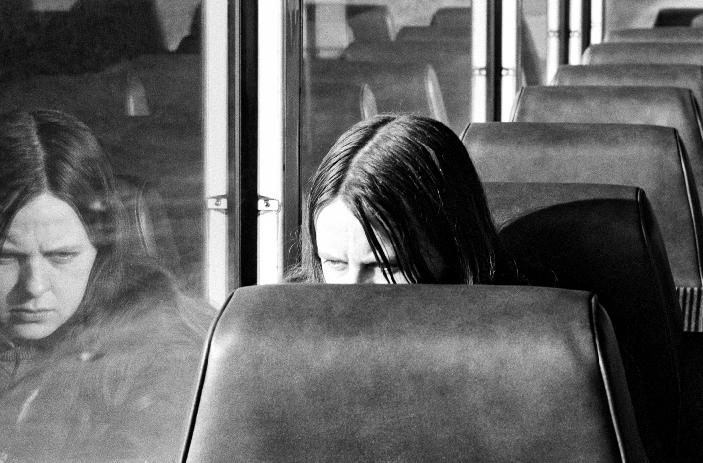A black and white image of a woman sitting alone in a row of seats, as if on a bus or train. Her face is obscured by the seat in front of her from the nose down, but her full reflection is visible in the window next to her.