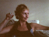 A grainy image of a woman looking to her left. She is leaning against the back of a chair, one arm outstretched while the other is bent and holds a cigarette.