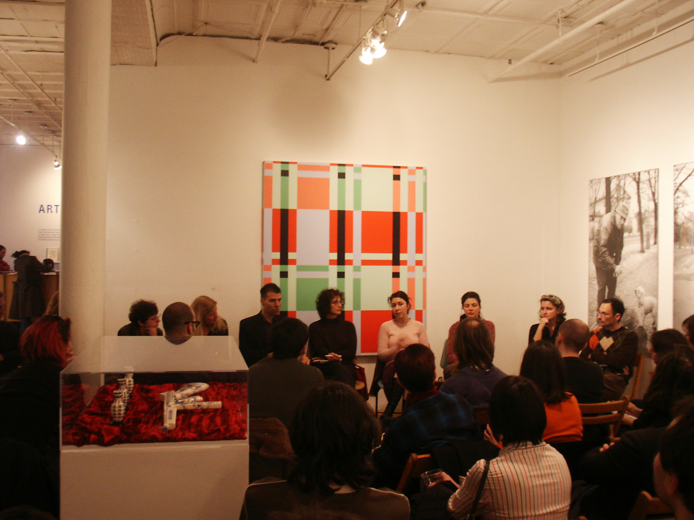 A circle of people sitting in chairs in discussion. Facing towards them are additional rows of chairs filled with an audience. To the left of the audience, there is a white plinth with a glass top housing several porcelin objects on a red fabric. On the wall behind the circle of speakers, there is a plaid composition featuring red, green, and peach colors.