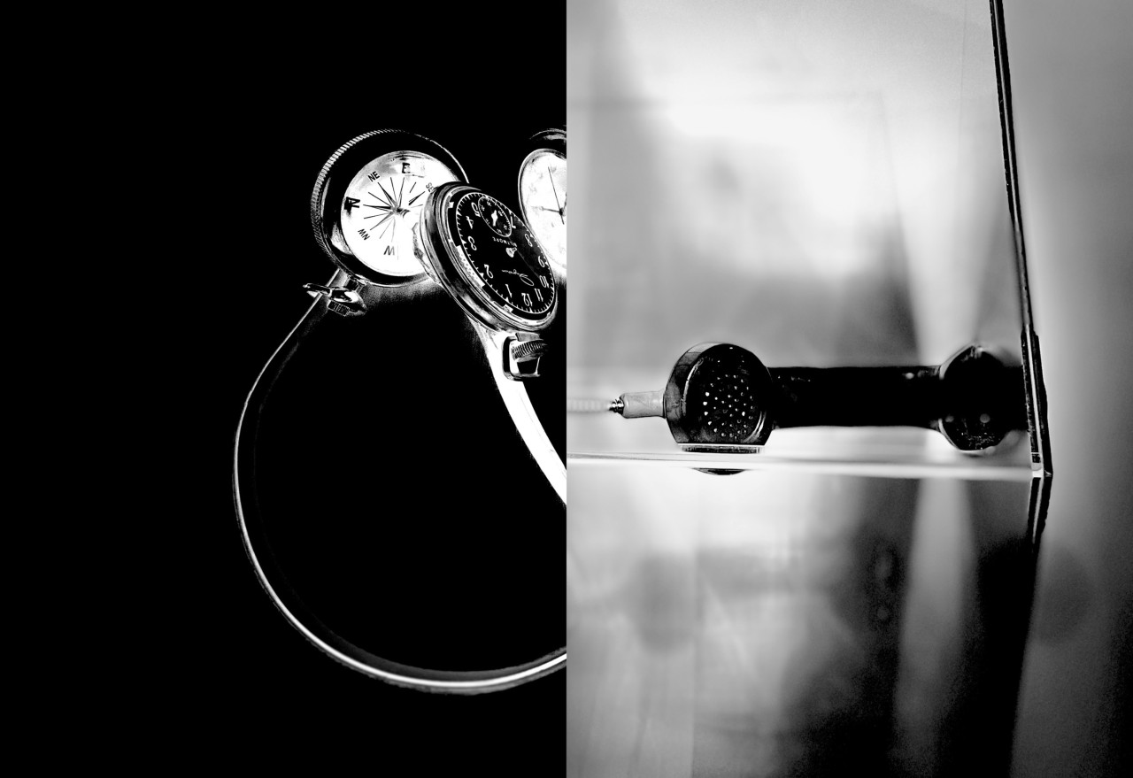 Two photographs are shown in a book spread. On the left page, a high-contrast black-and-white photograph hosts heavy light and shadow play. An iconic payphone handset is centered floating in a clear cube, with its top protruding slightly above the cube’s top surface. The telephone’s armored cord hangs down in loose loops inside the cube, its exposed wire slack touching the interior bottom. On the right page, a black-and-white photograph shows two pairs of modified headphones entwined on a deep black surface. In place of speakers, one headphone strap hosts clocks that each have a small stopwatch dial; the other strap hosts a pocket watch and a compass. The clock hands are missing from the clocks; inside their larger clockface circles, each clock hosts a smaller circle with a stopwatch.