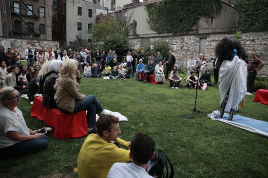 A stone-walled lawn filled with people watching Kembra Pfahler.