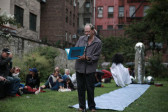 Man standing outside at a microphone in front of a large silver inflatable penis, reading from a purple folder.