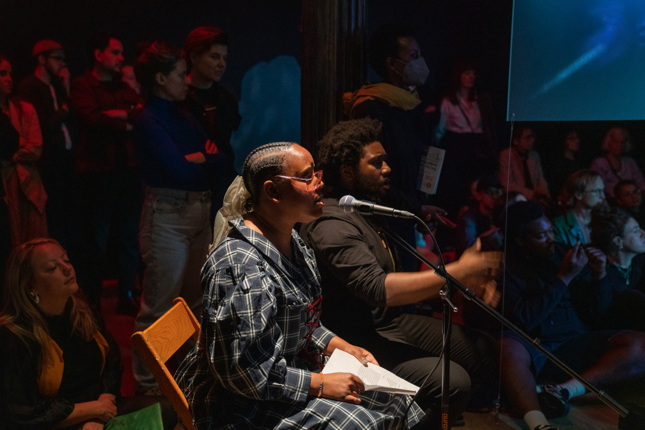Color photograph of two performers, seated side by side, singing into a microphone stand. Next to them can be seen the corner of a hanging screen positioned in the center of the room. Behind and around the performers audience members sit on the floor or stand.
