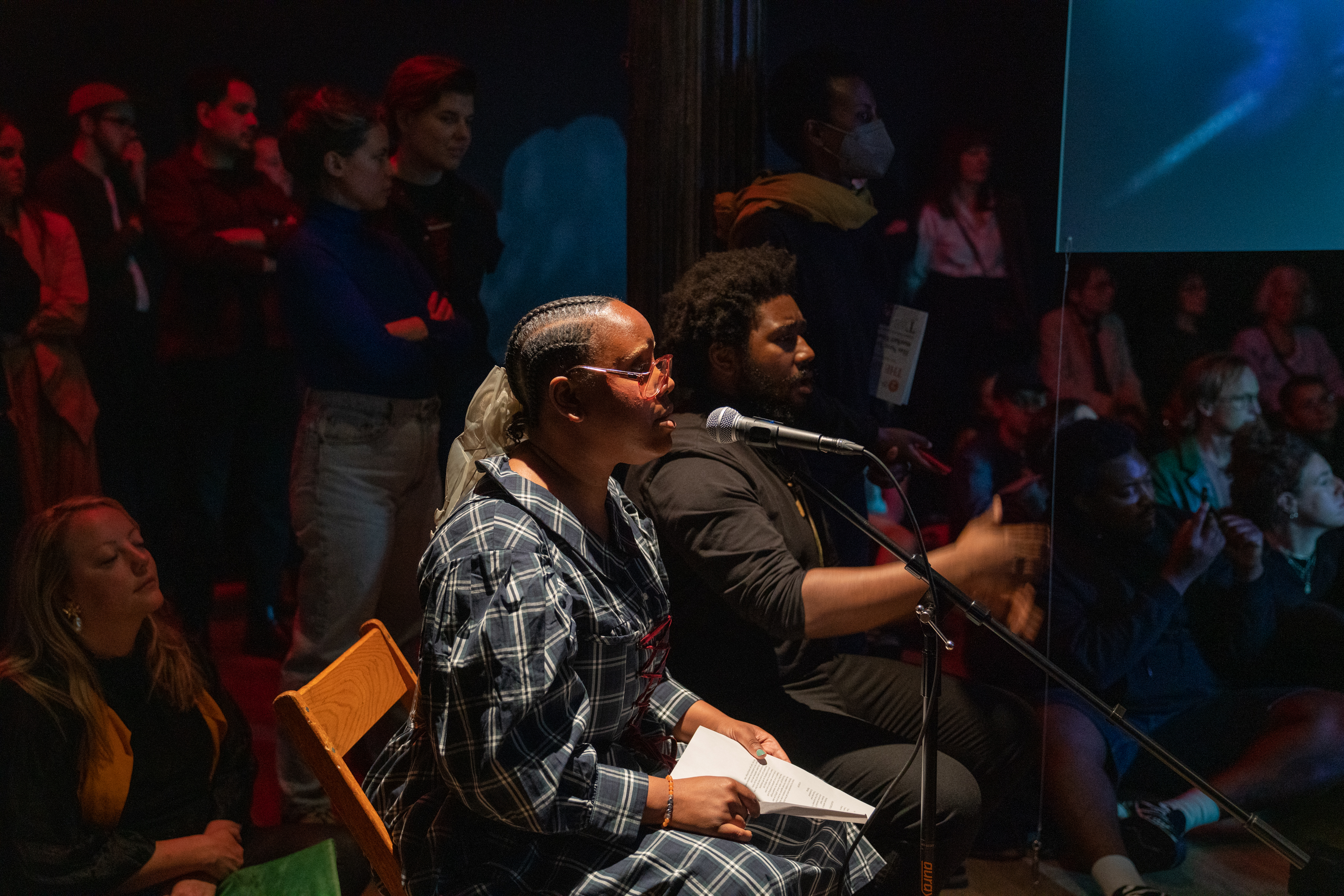 Color photograph of two performers, seated side by side, singing into a microphone stand. Next to them can be seen the corner of a hanging screen positioned in the center of the room. Behind and around the performers audience members sit on the floor or stand.
