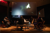 Color photograph of a group of performers, seated in a circle around a gallery room. The performers include a choruse of singers, a pianist, and a harpist with their instruments. They sit underneath three hanging screens that surround the center of the room. The screens depict a video still of a figure's face, obscured by a bright light in one corner.