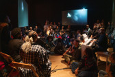 Color photograph of the center of a gallery room, in which performers are seated in wooden chairs around the center, alongside three hanging screens that project a video still of a figure's face against a dark background. All around the room, audience members sit on the floor or stand.