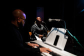 Color photograph of two figures preparing to perform. In the foreground, out of focus, is the side profile of a figure playing a white keyboard, with music sheets in front of them. To the right, in focus, another figure sits on a wooden chair, eyes closed in concentration and behind a microphone stand.