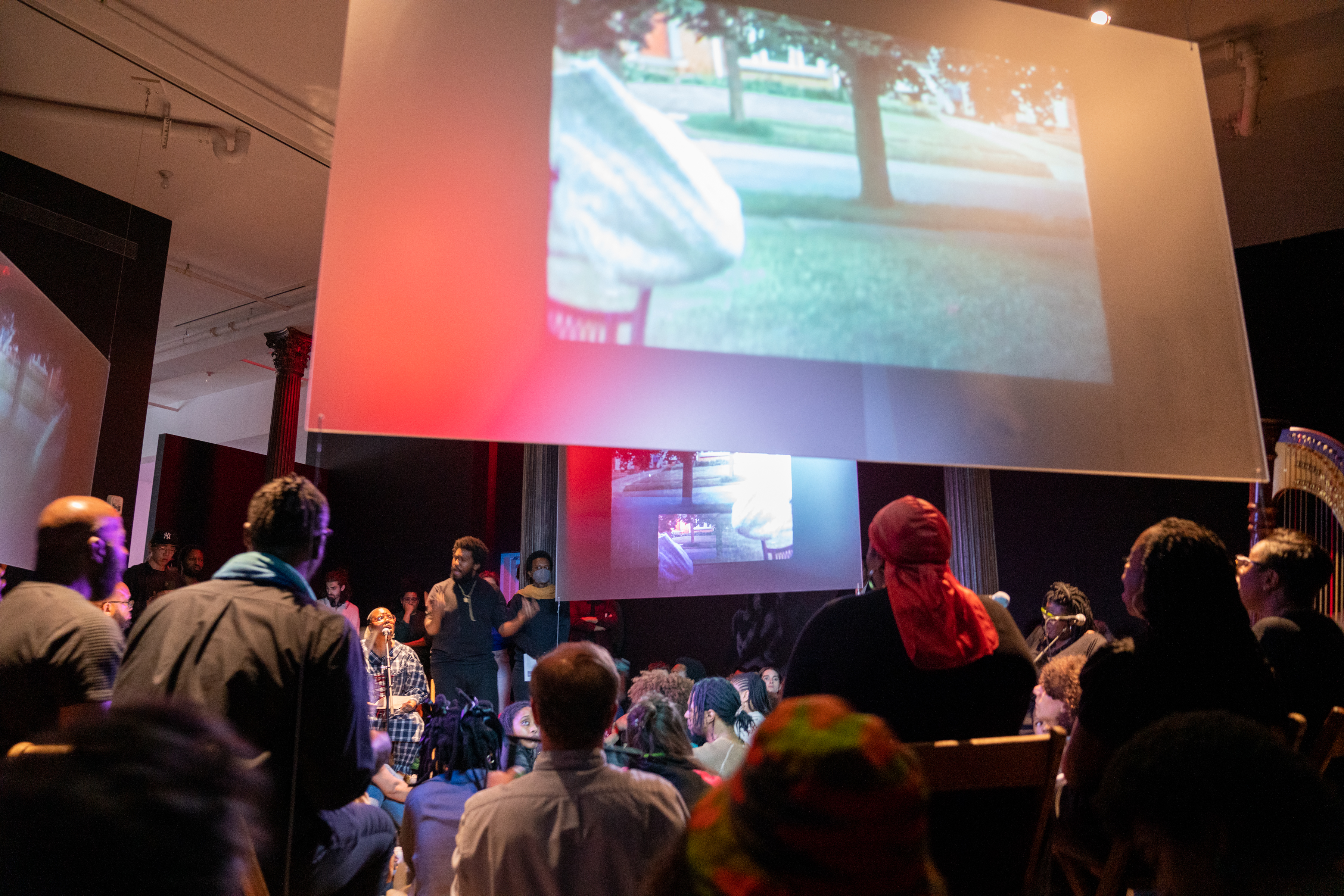 Color photograph, taken from a low angle, looking up at three hanging screens positioned around the center of a gallery room. Below the screens, performers are seated in wooden chairs, singing into microphones. A performer in the background stands and waves his hands to conduct the group. All around the performers, audience members fill the room.