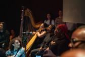 Color photograph of a harpist, and a row of singers seated, and singing into a microphone stand. Audience members fill the room on either side of the seated performers.