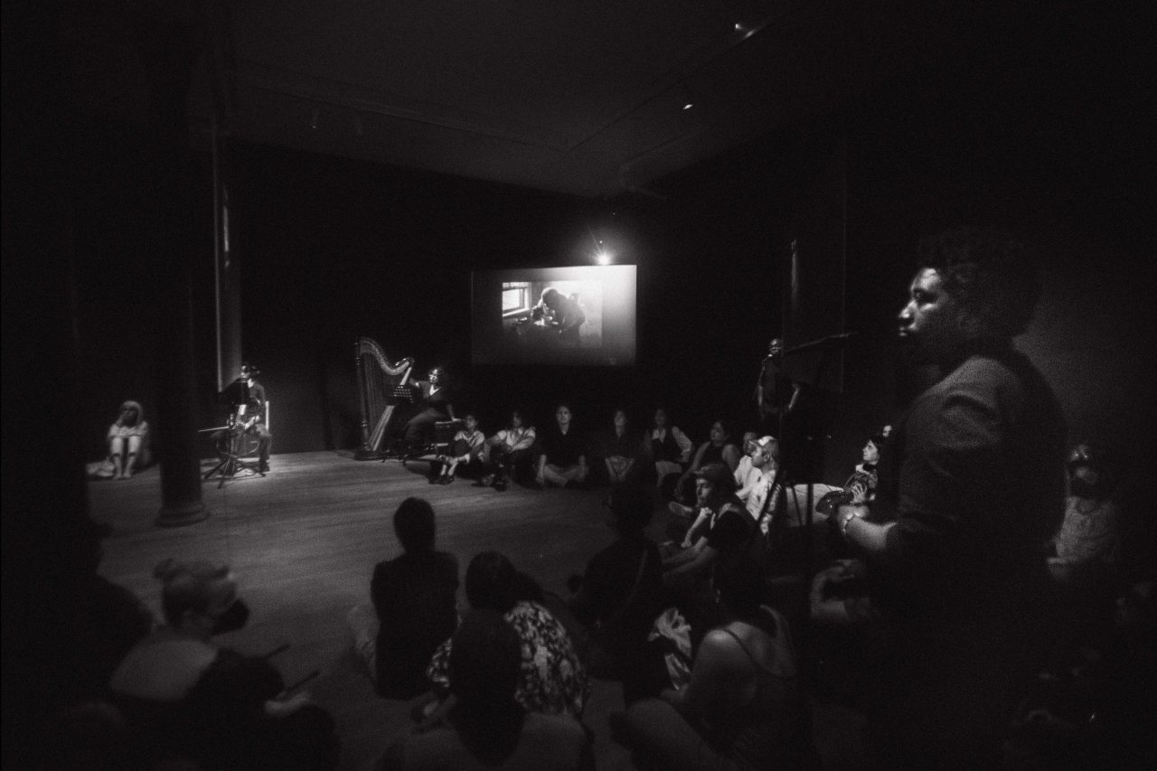 Black & white photograph of a gallery room with black walls. Around the center of the room are three hanging screens. Around the edges of the room, performers wearing all black stand under spotlights. Along the back wall sit a cellist and harpist with their instruments. The audience sit on the floor in front of the performers.