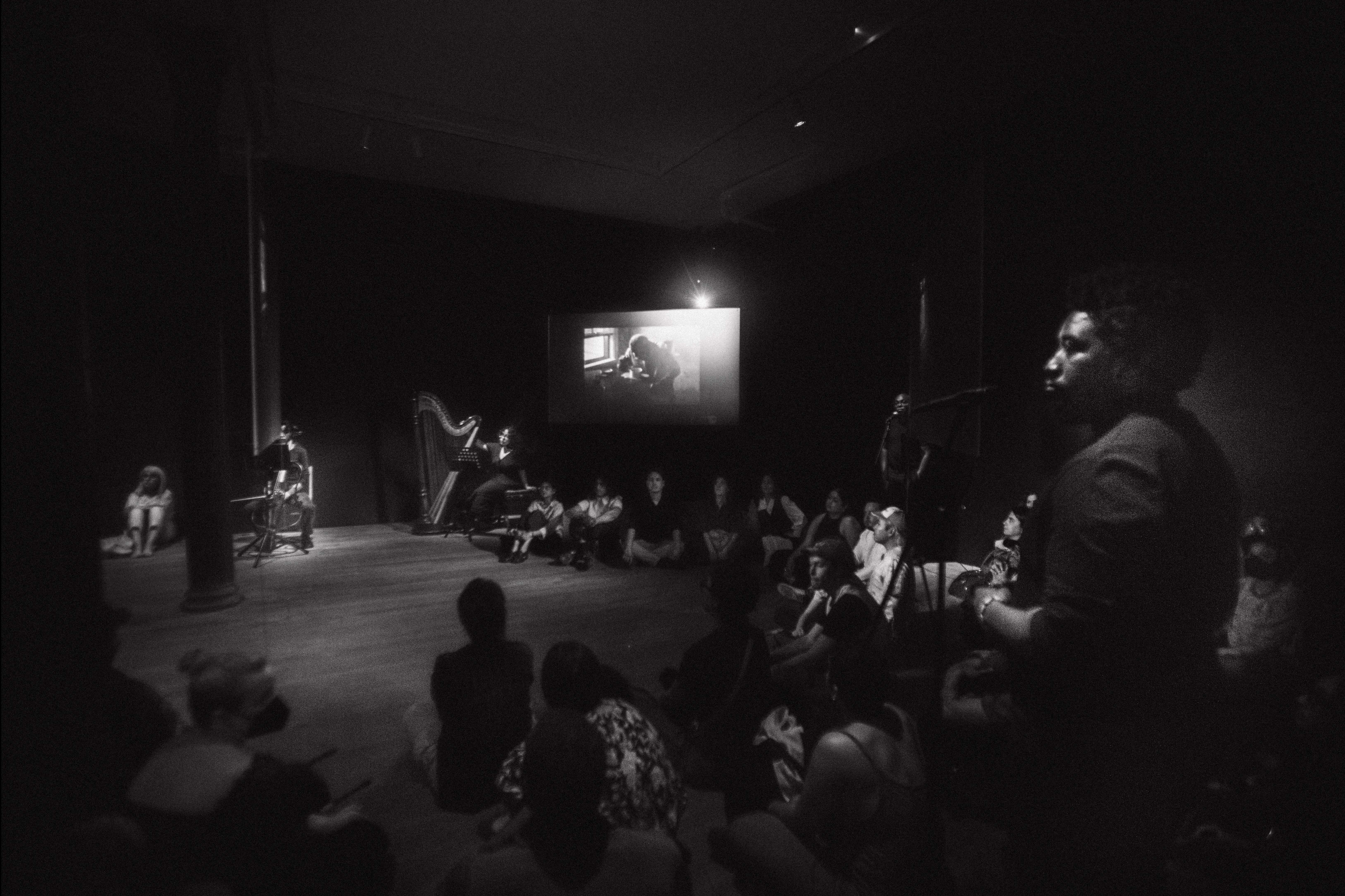 Black & white photograph of a gallery room with black walls. Around the center of the room are three hanging screens. Around the edges of the room, performers wearing all black stand under spotlights. Along the back wall sit a cellist and harpist with their instruments. The audience sit on the floor in front of the performers.