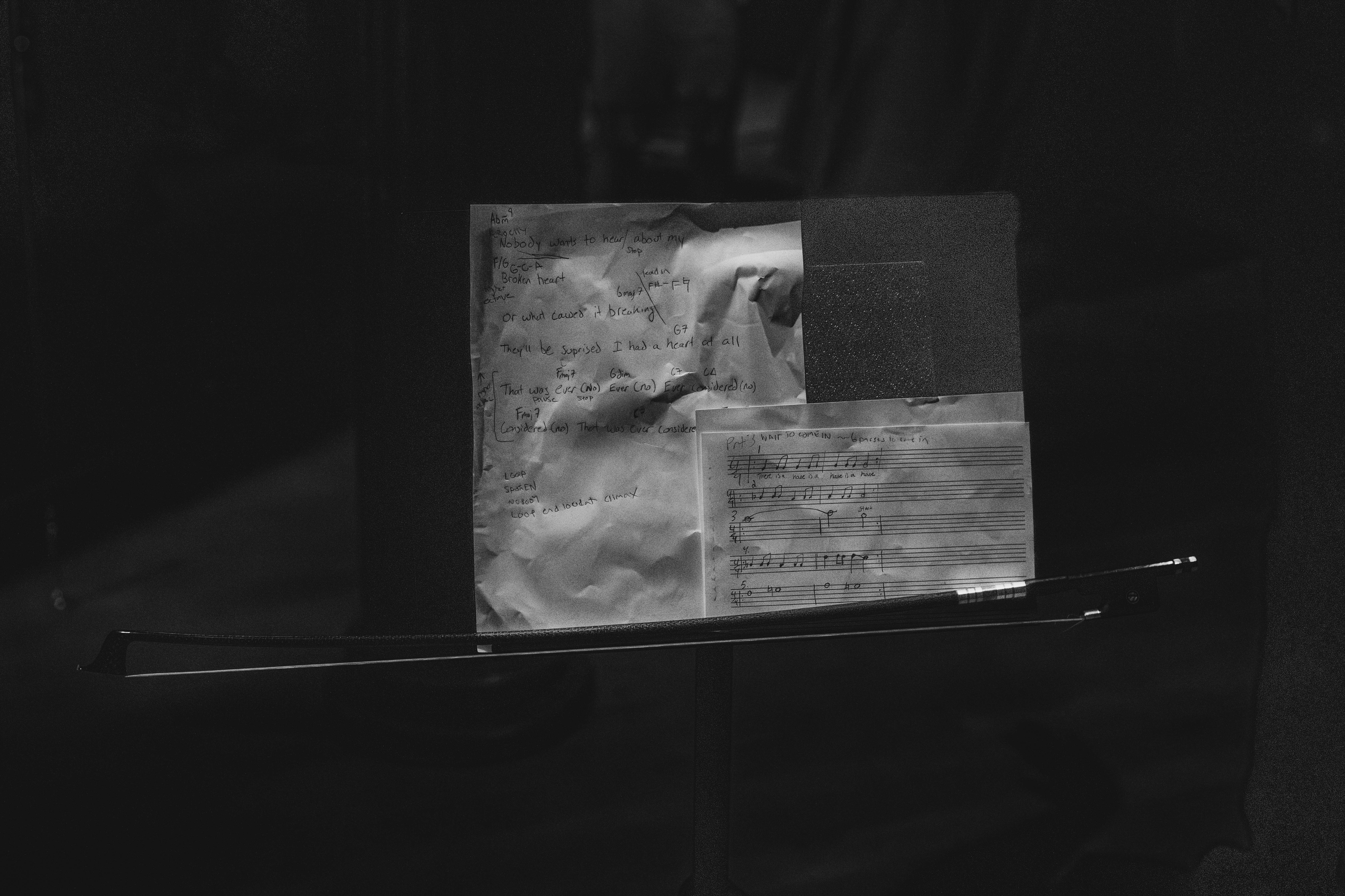 Black & white photograph of a music score and handwritten lyrics on a music stand, with a cello bow.