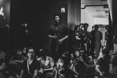 Black & white photograph of one side and the entrance into a gallery room. The walls are painted black, and at the corner of the entrance stands a performer singing into a microphone stand. Around him and along the wall audience members sit on the floor or stand.