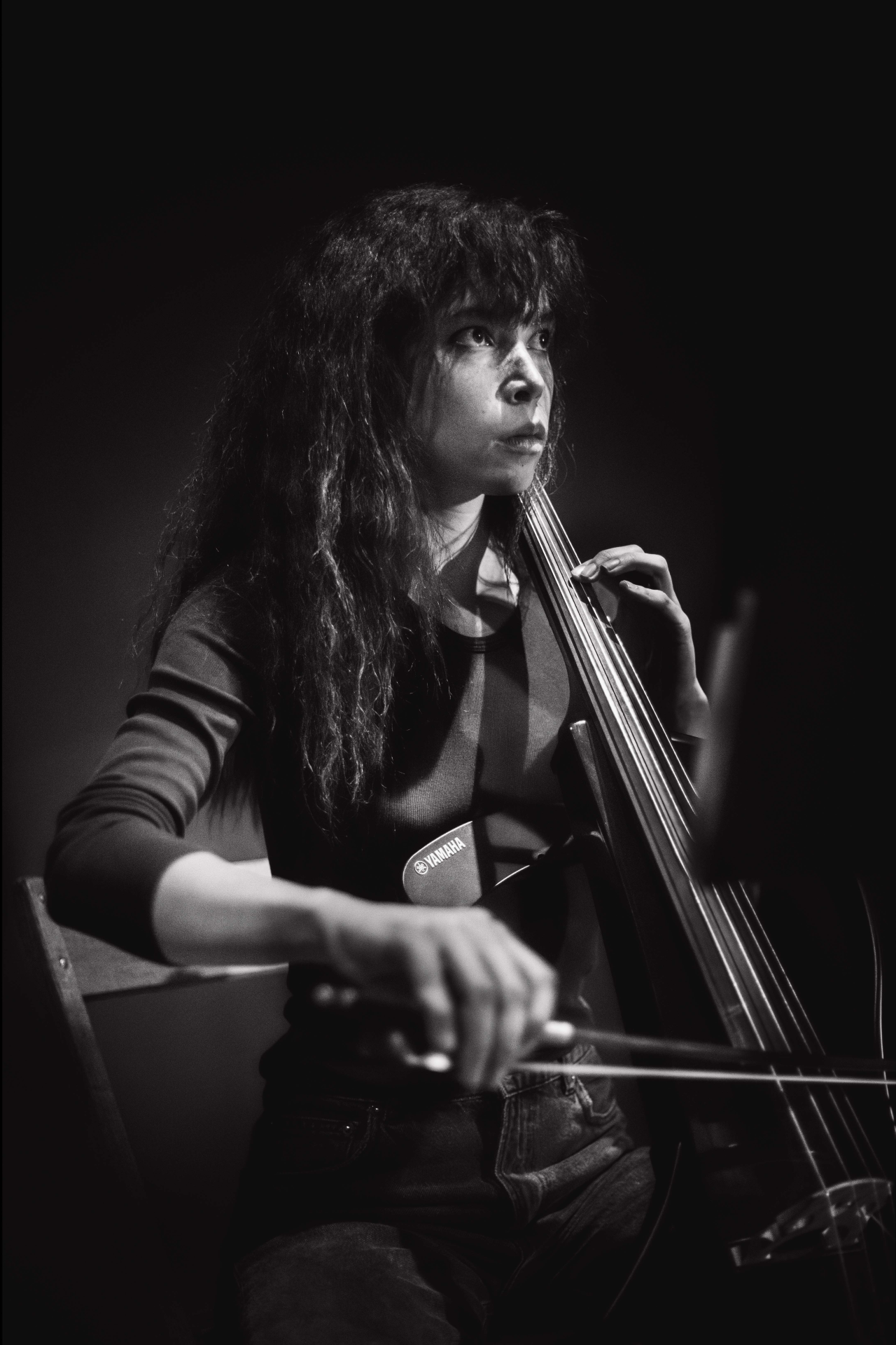 Black & white photograph of a performer sitting in a wooden chair. A spotlight shines on them from above, and they play a cello that leans against their body.