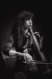 Black & white photograph of a performer sitting in a wooden chair. A spotlight shines on them from above, and they play a cello that leans against their body.