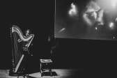 Black & white photograph of a room with black walls. A figure stands behind a harp, next to a stool and music stand. A spotlight shines on the harp. In the middle of the space is a hanging screen that plays a projected video of a close-up of a face.