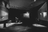 Black & white photograph of an empty gallery room, with black walls. Hanging around the center of the space are three hanging screens playing the same synchronized video. Along the back wall a cello lays against a wooden chair, next to a harp and stool. On the side wall a spotlight shines on a microphone stand.