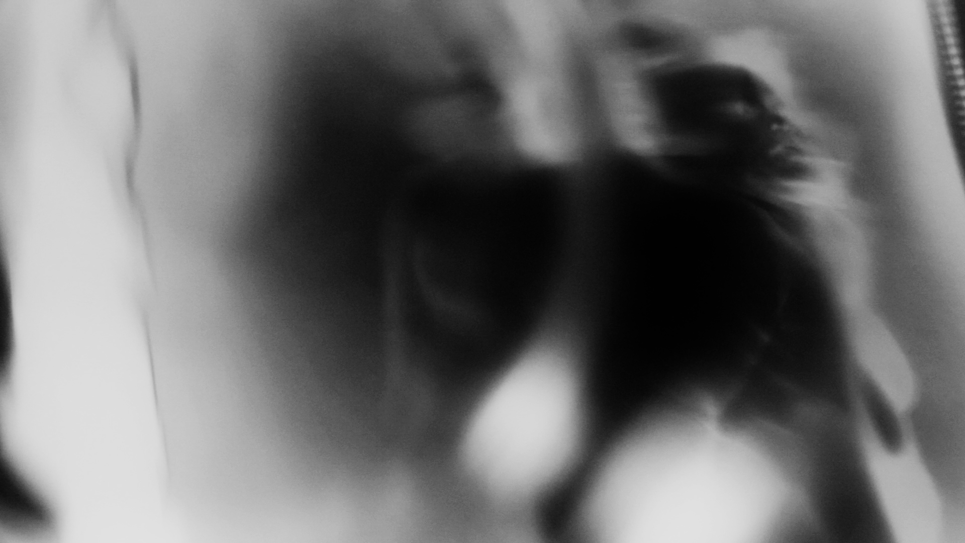 A black-and-white film still of hazy and ghostly tonal modulations. Within the tonal atmosphere, fragments of a person's face and body can be made out in the right half of the image.