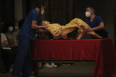 Two figures wearing medical scrubs hold a mannequin over a table.