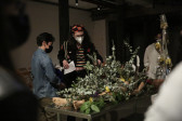 Various figures stand around a table with a mannequin on it. The mannequin is obscured by foliage and flowers.