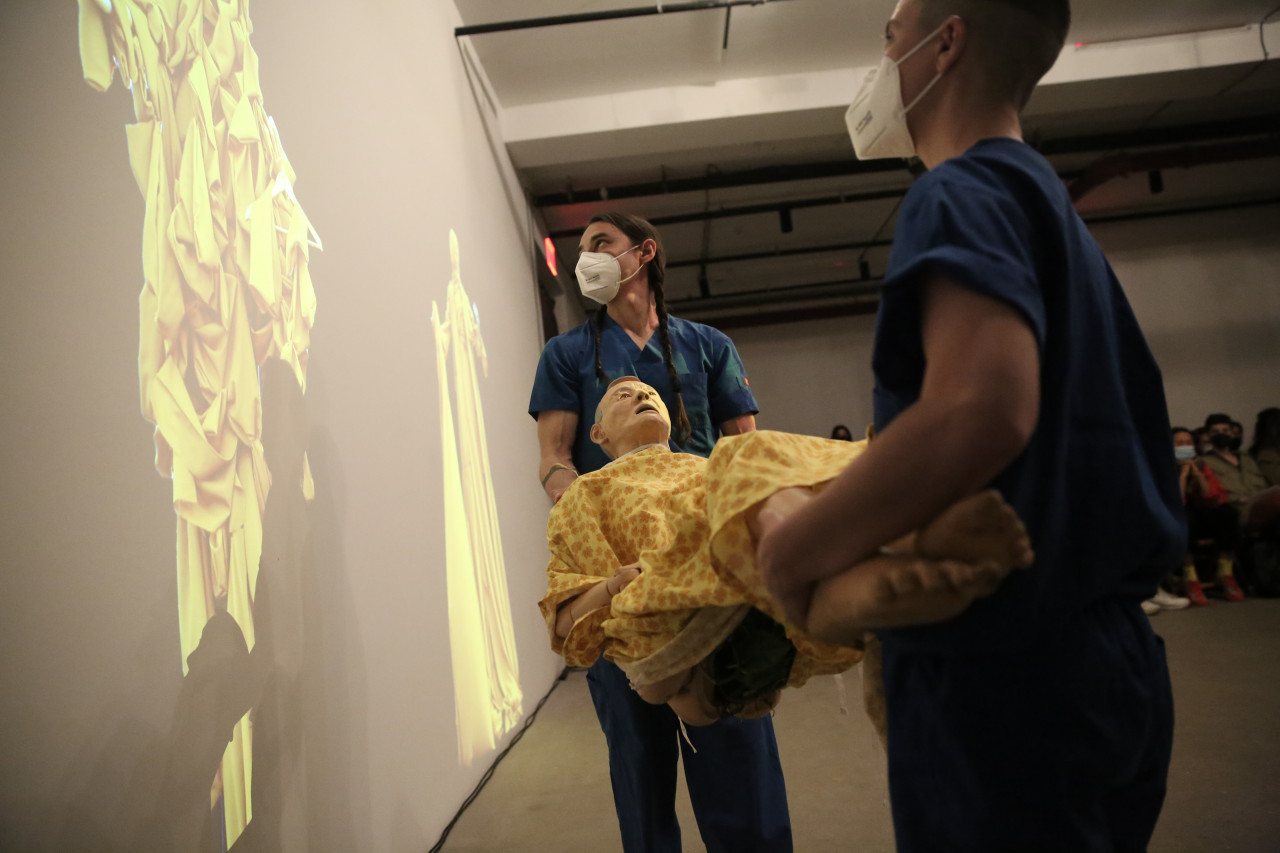 Two figures holding a mannequin look at a projection on the wall.
