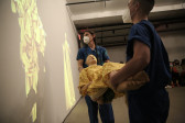 Two figures holding a mannequin look at a projection on the wall.