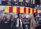 Photograph of two figures, one in a suit, one in a suit and brown trenchcoat, striding across a city street. The facade of the buildings behind them is covered with a colorful mural and a long line of alternating, bright red and yellow posters. A large building sign above the posters reads 