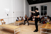 A man stands, motioning towards at an LED light sculpture hung on the wall. A woman sits on one of three benches circling the man, and the audience is seated on the floor behind them.