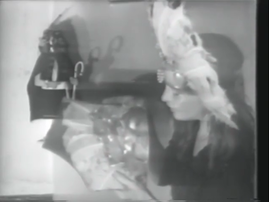 A blurry black-and-white photo of someone wearing a large white headdress holding several unidentifiable objects.