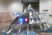 Sculpture of an alien spaceship made out of sheet metal, with a red and blue LED light, sitting in a gallery.