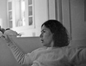 A black and white image of a person with dark hair looking to the left with their arms outstretched. In the distance is a dish rack with dishes and a window flanked by cabinets.