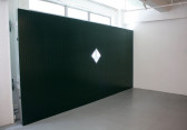 Installation view of a mechanical wall covering a window in a gallery space, with a diamond-shaped glass pane in the center letting light through.