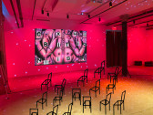 A basement gallery is filled with red light and disco lights. On the far wall is projected a book cover featuring a painting of a naked body with body parts squished together. Fake chairs drawn in black outline are placed around the space.